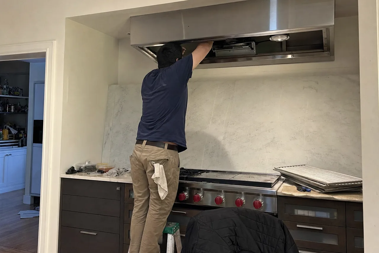 A technician on a small step stool reaches up into a stainless wall hood with its filters out, above a rangetop with red knobs and a griddle, the baffle filters stacked on the counter
