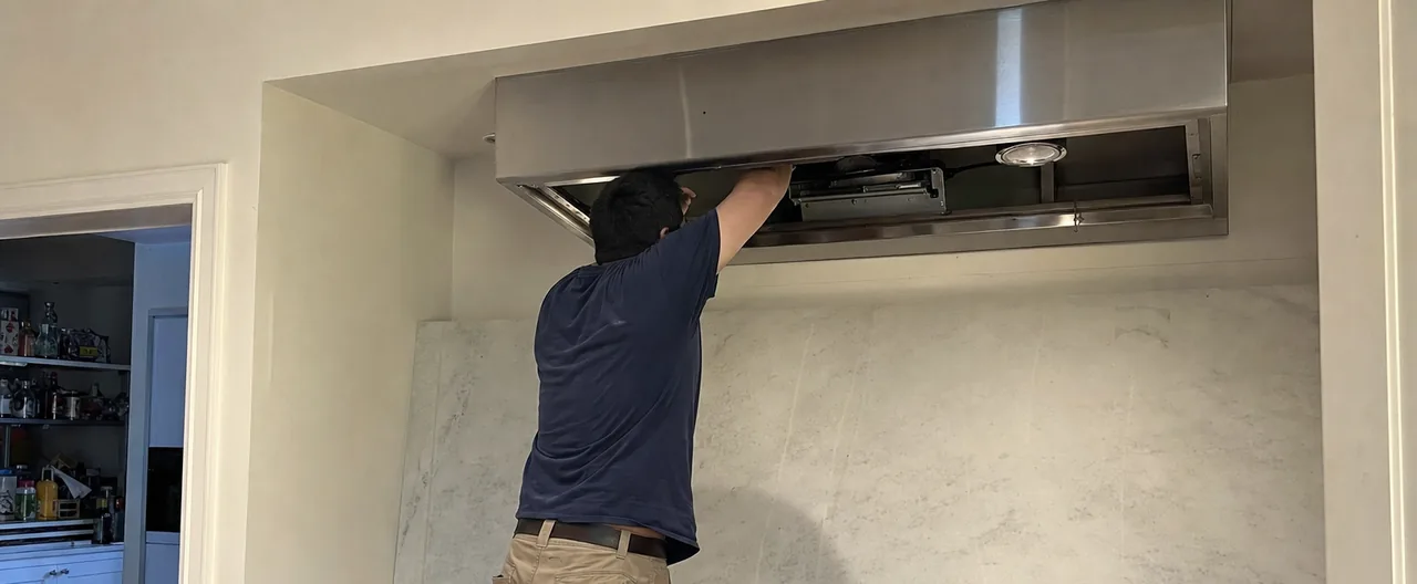 A technician on a step stool reaching up into an open stainless hood canopy, baffle filters stacked on the counter